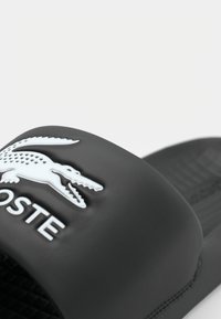 Sort gummisandal med struktureret sål, med et hvidt hævet krokodillelogo og "LACOSTE" branding på remmen.