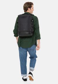Sac à dos noir avec des sangles réglables, de forme rectangulaire, en tissu texturé, posé sur une personne portant un jean retroussé et des baskets.