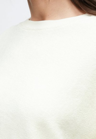 Φόρμα σε ανοιχτό μπεζ χρώμα με υφή υφάσματος. Διαθέτει ριμπ λαιμόκοψη και μαλακή, άνετη εφαρμογή. Κοντινή όψη που δείχνει λεπτομέρειες και υλικό.