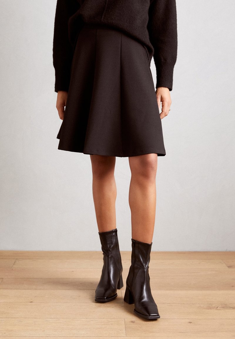 Černá rozšířená sukně vyrobená z texturované tkaniny, v kombinaci s lesklými černými kotníkovými botami na blocím podpatku, umístěné na světlém pozadí.