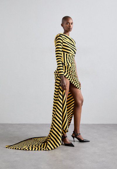 Randig långärmad klänning i gult och svart med en dramatisk hög-låg fåll och rynkade detaljer, tillsammans med svarta stilettklackar.