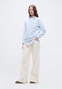 Νεαρή γυναίκα στέκεται με το χέρι στην τσέπη, φορώντας ανοιχτό μπλε φούτερ πάνω από ριγέ πουκάμισο, φαρδιά λευκά παντελόνια και σκούρα παπούτσια σε απλό φόντο.