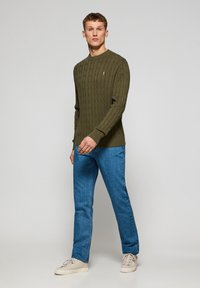 Khaki cable-knit sweater with a rounded neckline, paired with light blue denim jeans and beige sneakers.