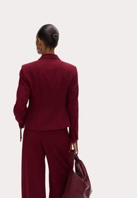 Chaqueta de traje burdeos con un diseño estructurado, que presenta un cuello con muescas y detalles de botones, acompañada de pantalones de pierna ancha a juego.