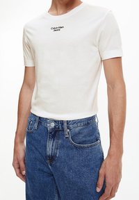 Bílý bavlněný tričko s krátkým rukávem a kulatým výstřihem, s černým logem "Calvin Klein Jeans" na hrudi. V kombinaci s modrými džíny.