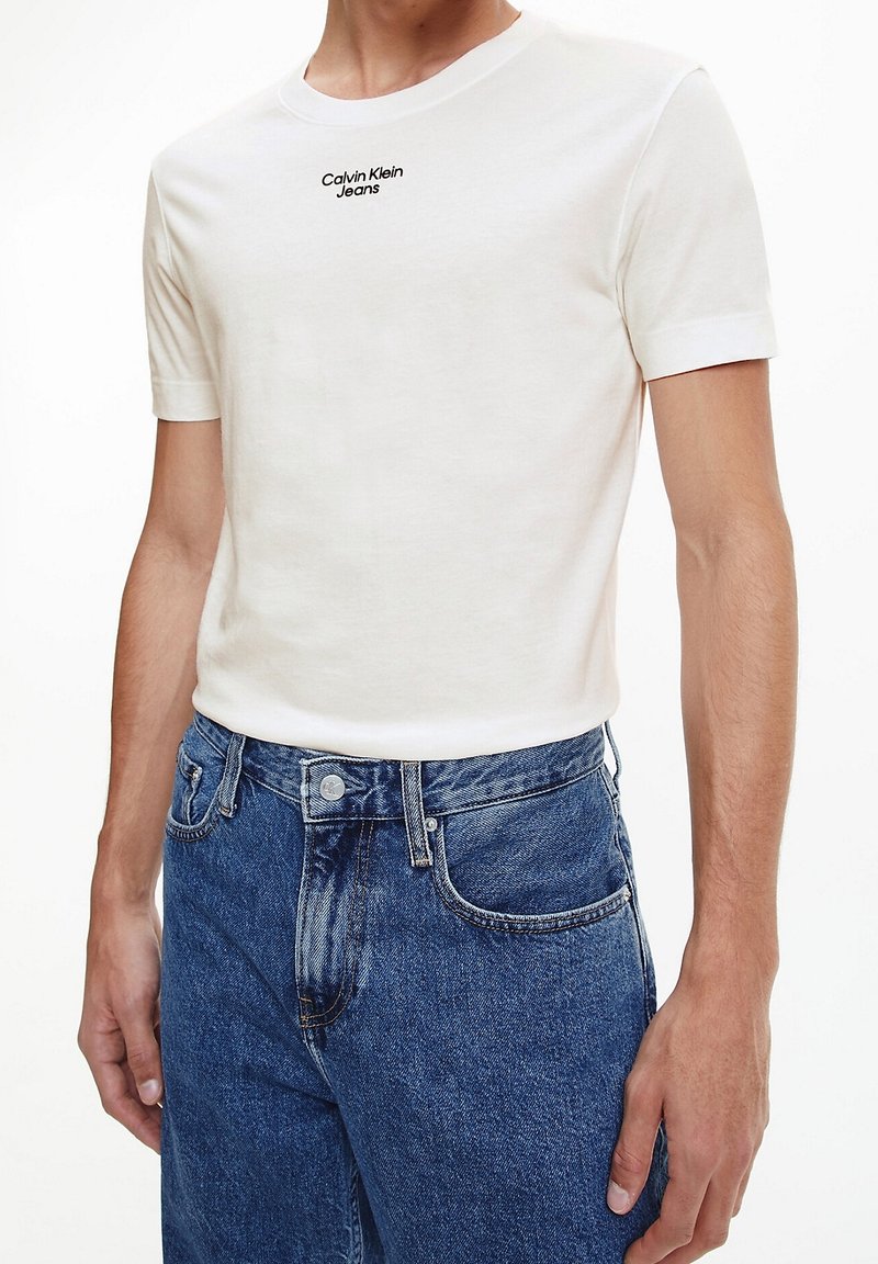 Bílý bavlněný tričko s krátkým rukávem a kulatým výstřihem, s černým logem "Calvin Klein Jeans" na hrudi. V kombinaci s modrými džíny.