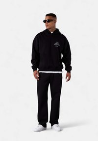 Haut à capuche noir avec une poche avant et un logo blanc, associé à un pantalon de survêtement noir assorti et des baskets blanches. Le modèle porte des lunettes de soleil.