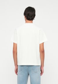 Mężczyzna z ciemnymi włosami ubrany w prostą białą koszulkę z krótkim rękawem i jasnoniebieskie dżinsy, stojący przodem do białego tła.