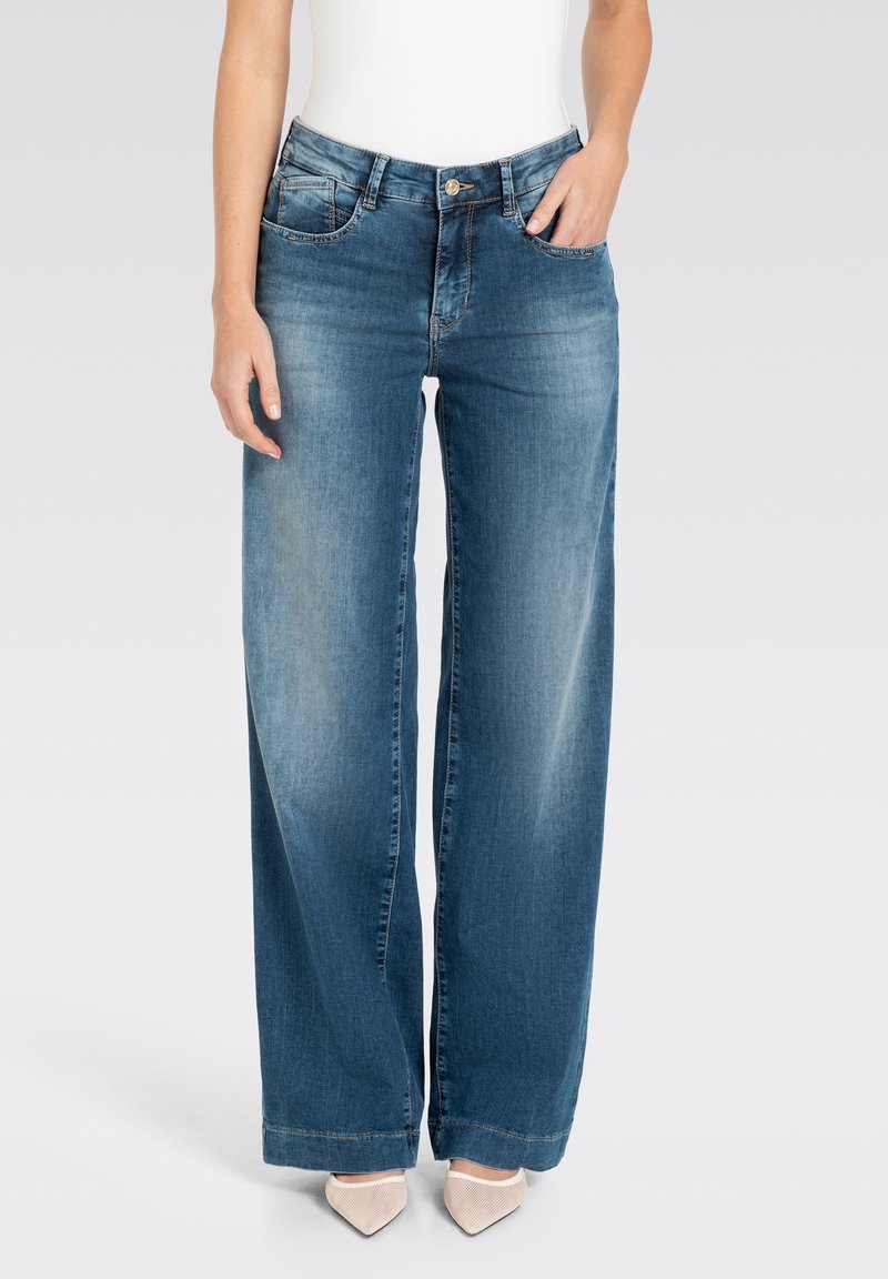 Vrouw draagt medium-wash jeans met wijde pijpen en vervaging aan de voorkant van de dijen, een wit shirt in de broek gestopt en beige schoenen met puntige neus, staand tegen een effen achtergrond.