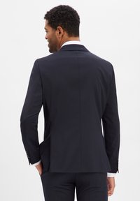 Marineblaue Anzugjacke aus glattem Stoff, maßgeschneiderte Passform, ein Schlitz, weiße Hemdmanschetten sichtbar, polierte Knöpfe und leicht strukturierte Schultern.