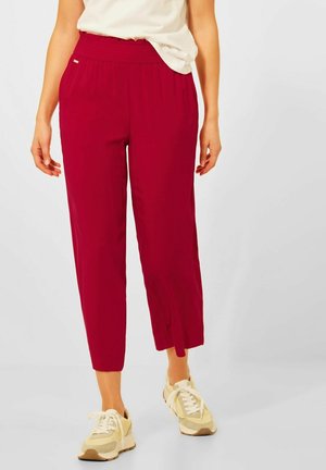 Femme portant un pantalon rouge court avec des fentes à l'avant et des baskets beiges, debout devant un fond uni.
