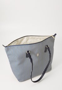Sac fourre-tout rayé en marine et blanc, fabriqué en tissu à la texture lisse. Il possède des poignées doubles et un logo doré à l'avant.