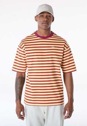 Junger Mann, der ein rot-gelb-weiß gestreiftes T-Shirt, hellgraue Hose, eine hellgraue Kappe trägt, mit Tattoos auf seinem linken Unterarm.