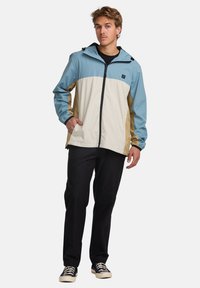 Lightweight, multi-colored jacket featuring a blue top, cream middle, and tan sides, with a zipper and adjustable hood. Worn with black pants.
