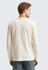 Koszula z długim rękawem w jasnym kremowym kolorze, wykonana z miękkiej tkaniny, z prostym dekoltem i prostym krojem, przedstawiona od tyłu.