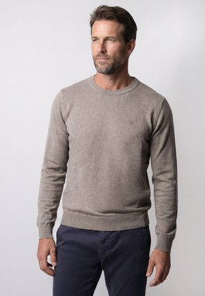 Man draagt een beige trui zonder kraag en donkere broek, staat tegen een rustige lichte achtergrond en kijkt iets naar rechts.