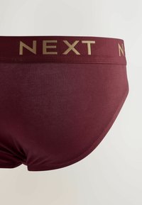 Culottes en coton bordeaux avec un logo doré "NEXT" sur la ceinture. Le tissu semble lisse avec une coupe ajustée et sans motifs visibles.