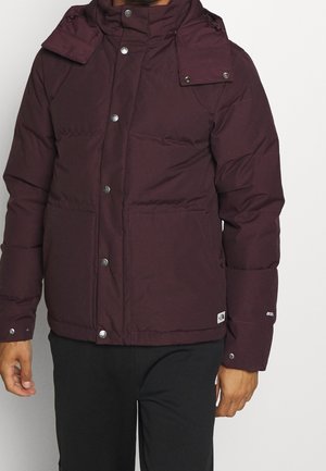 Mann trägt eine dunkel kastanienrote gesteppte Winterjacke mit Knopf- und Reißverschluss sowie schwarze Hose vor einfarbigem Hintergrund.