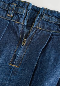 Pantalon en denim bleu avec fermeture éclair, présentant des accents cousus et un design plissé. Le tissu affiche une texture en denim classique.