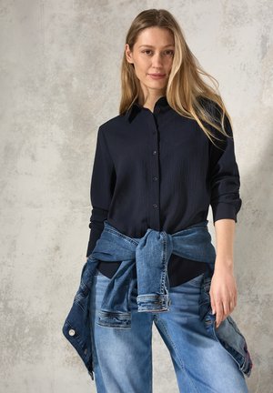Marineblauwe overhemdbloes met een gestructureerde afwerking, gecombineerd met lichte spijkerbroek en een vastgebonden spijkerjack om de taille.