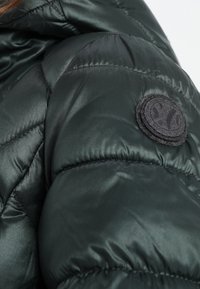 Manga de chaqueta acolchada de color verde oscuro con textura acolchada; presenta un parche negro redondo con diseño en relieve. Material liso y brillante.