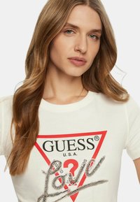 Weißes T-Shirt mit Rundhalsausschnitt, das ein rotes, dreieckiges Logo mit dem aufgedruckten Text "GUESS" und "U.S.A." zeigt; verziert mit einem silbernen, mit Schmuck besetzten Schriftzug "Love".
