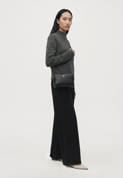 Une femme présente un pull gris anthracite, un pantalon noir à jambes larges et un petit sac bandoulière noir sur un fond clair uni.