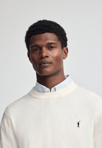 Cremfarbener Strickpullover mit Rundhalsausschnitt, versehen mit einem kleinen gestickten Logo. Über einem hellblauen und weißen gestreiften Hemd mit Kragen getragen.