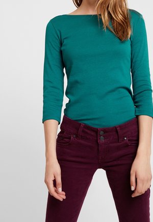 Vrouw draagt een turquoise top met lange mouwen en een donkerpaarse broek met hoge taille, staat met ontspannen armen langs haar lichaam tegen een effen achtergrond.