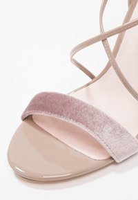 Sandale beige à bout ouvert avec une lanière avant en velours rose pâle et de fines lanières beiges à la cheville sur fond blanc.