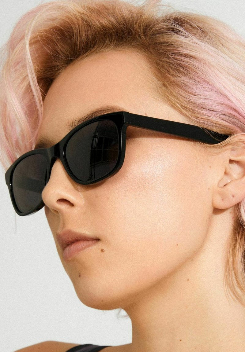 Black sunglasses with a bold, angular frame and dark lenses. Features thick temples and matte finish. Hair has soft pink highlights.