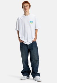 Weißes Baumwoll-T-Shirt mit kurzen Ärmeln, das ein blau-grünes Grafikmotiv auf der Brust zeigt. Kombiniert mit weiten blauen Denim-Jeans und weißen Sneakers.