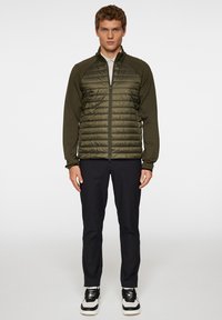 Nuori mies seisoo kameran edessä, päällään oliivinvihreä vetoketjuhuppari ja mustat housut, jalassaan valkoiset lenkkarit yksinkertaisella taustalla.