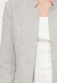 Ανοιχτό γκρι σακάκι με σχέδιο ψαροκόκκαλο, καλοσχηματισμένο γιακά και εφαρμοστή γραμμή. Φορεμένο πάνω από λευκό μπλουζάκι με κάθετες ραβδώσεις.