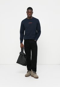 Navy long-sleeve shirt with red text, black pants, gray boots, holding a black quilted bag. Simple design, made from fabric.