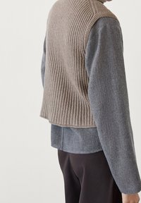 Vahva beige villatakki, joka on kerroksittain harmaan pitkähihaisen paidan päällä. Villatakissa on lyhyt helma ja teksturoitu neulekangas.