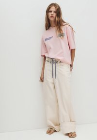 Lys pink bomulds T-shirt med blå tekst, parret med cremefarvede bukser med brede ben, som har en blå snøre og sølvfarvet cirkulært hardware.