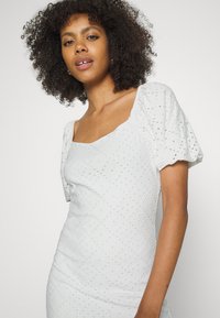 Vestido branco com decote quadrado, mangas curtas bufantes e tecido texturizado com padrões decorativos e recortes.