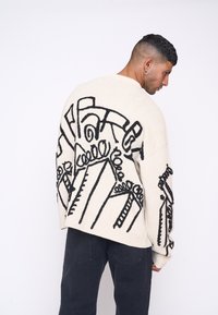 Pulover crem supradimensionat cu desene abstracte negre. Prezintă umeri căzuți și o textură moale, asortat cu blugi negri.