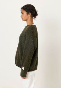 Žalias per didelis megztinis, pagamintas iš pūkuoto medžiagos, su apvalia iškirpte, nusileidusiomis petnešomis ir džersio kraštu. Ant rankovės yra etiketė.