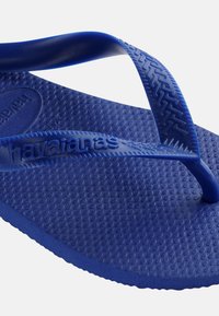 Suures plaanis sinine Havaianase plätu, millel on tekstureeritud tald ja nurga all olevatel kummirihmadel reljeefne kaubamärgi nimi.