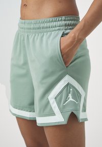 Close-up van een persoon die lichtgroene basketbalshorts met witte bies en Jordan-logo draagt, hand rustend in de zijzak.