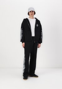 Svart hettegenser med sølv grafikkdetaljer på ermene, kombinert med matchende svarte joggebukser med et lignende design. Grå caps, svarte sneakers.