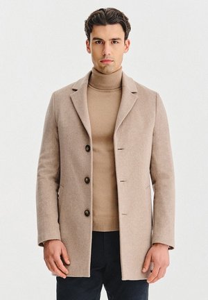 Beżowy wełniany płaszcz o strukturze z wyciętymi klapami i czterema guzikami. Stylizowany na beżowym golfie, uzupełniony ciemnymi spodniami.