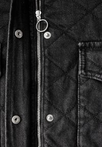 Chaqueta negra acolchada con una cremallera plateada, botones a presión y un tejido texturizado. Incluye un bolsillo en el pecho con costura visible.