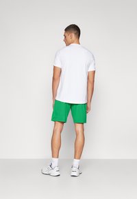 T-shirt bianco a maniche corte abbinato a pantaloni corti sportivi verdi, con un design testurizzato. Sneakers bianche e calzini alla caviglia completano il look.