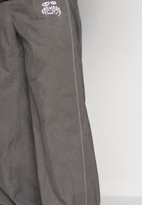 Gray cargo pants made from a lightweight, textured material, featuring white stitching and a prominent logo embroidered on the leg.