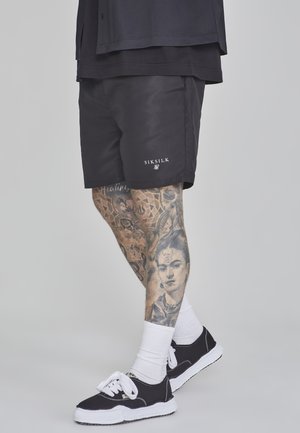 Pantaloni corti neri con logo SIKSILK, tessuto testurizzato, abbinati a calzini bianchi e sneakers neri. Tatuaggi sulle gambe ben visibili.
