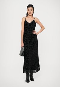 Abito maxi nero con sovrapposizione in pizzo floreale, spalline sottili, scollatura a V, silhouette fluida, abbinato a una borsa nera e stivali alti fino al ginocchio.