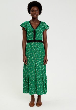 Kvinde stående iført en grøn blomstret V-hals kjole med sort kant og flagrende ærmer, kombineret med brune sandaler med åbne tæer.